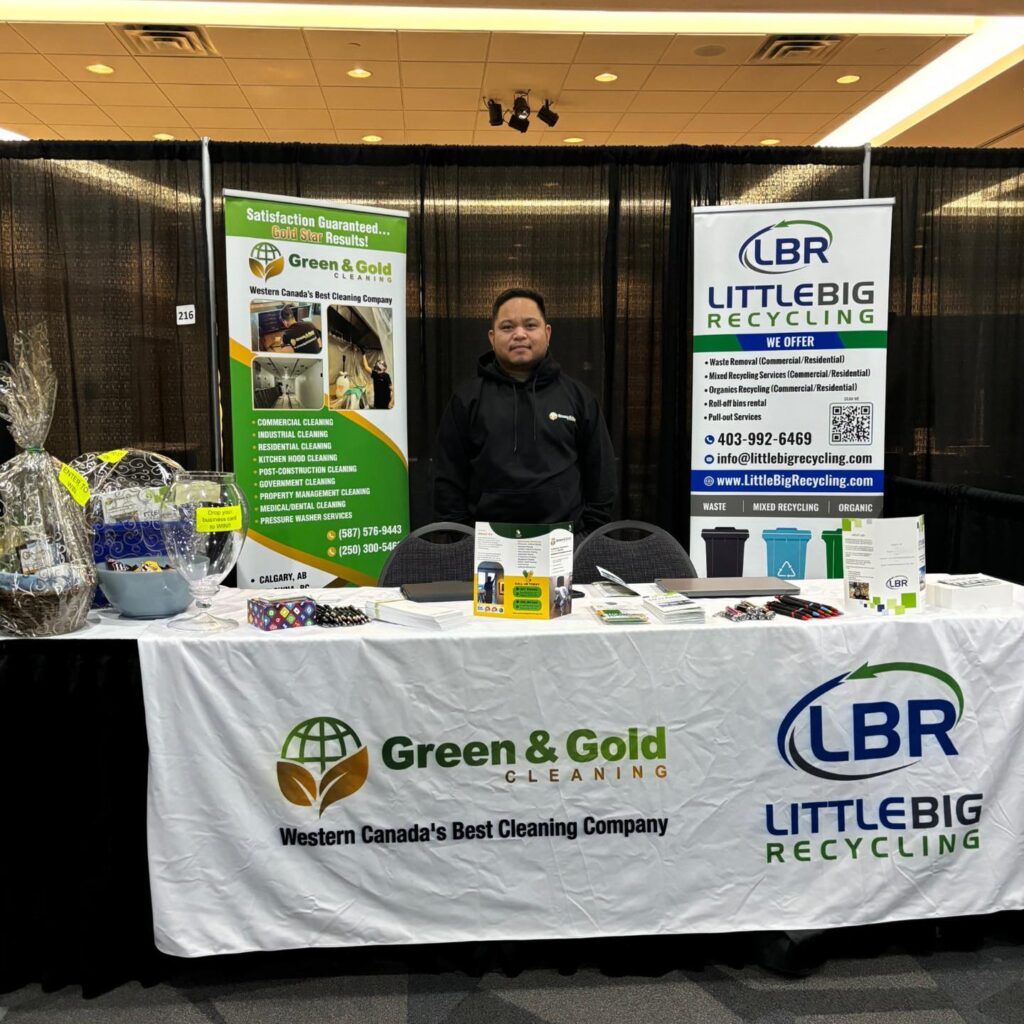 A man stands behind a promotional booth at an indoor event. The booth is for 'Clean It All' and 'LITTLEBIG RECYCLING.' Banners display services like commercial and residential cleaning, waste removal, and recycling. The tablecloth has the 'Clean It All' logo, which states 'Western Canada's Best Cleaning Company' and the 'LITTLEBIG RECYCLING' logo. On the table, there are business cards, informational pamphlets, and promotional giveaways, including a glass bowl filled with blue and white marbles and another with chocolate eggs wrapped in colorful foil.