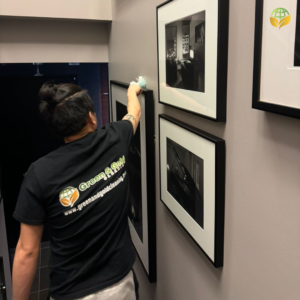 A person in a black shirt with the "Clean It All" logo cleaning a picture frame, demonstrating professional cleaning services' attention to detail.