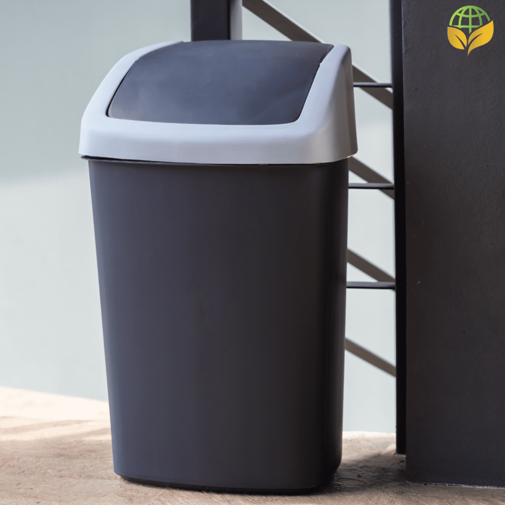 A black garbage can with a swinging lid, positioned next to a black metal fence on a sunlit patio. The can is made of plastic with a contrasting gray lid, and the background features a textured wall, partially shaded.