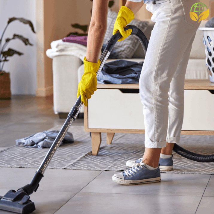 A person in casual attire with yellow rubber gloves is vacuuming a gray carpet in a living room setting. They are using a black vacuum cleaner. Visible in the background are a white sofa with purple cushions and a wicker basket with houseplants.